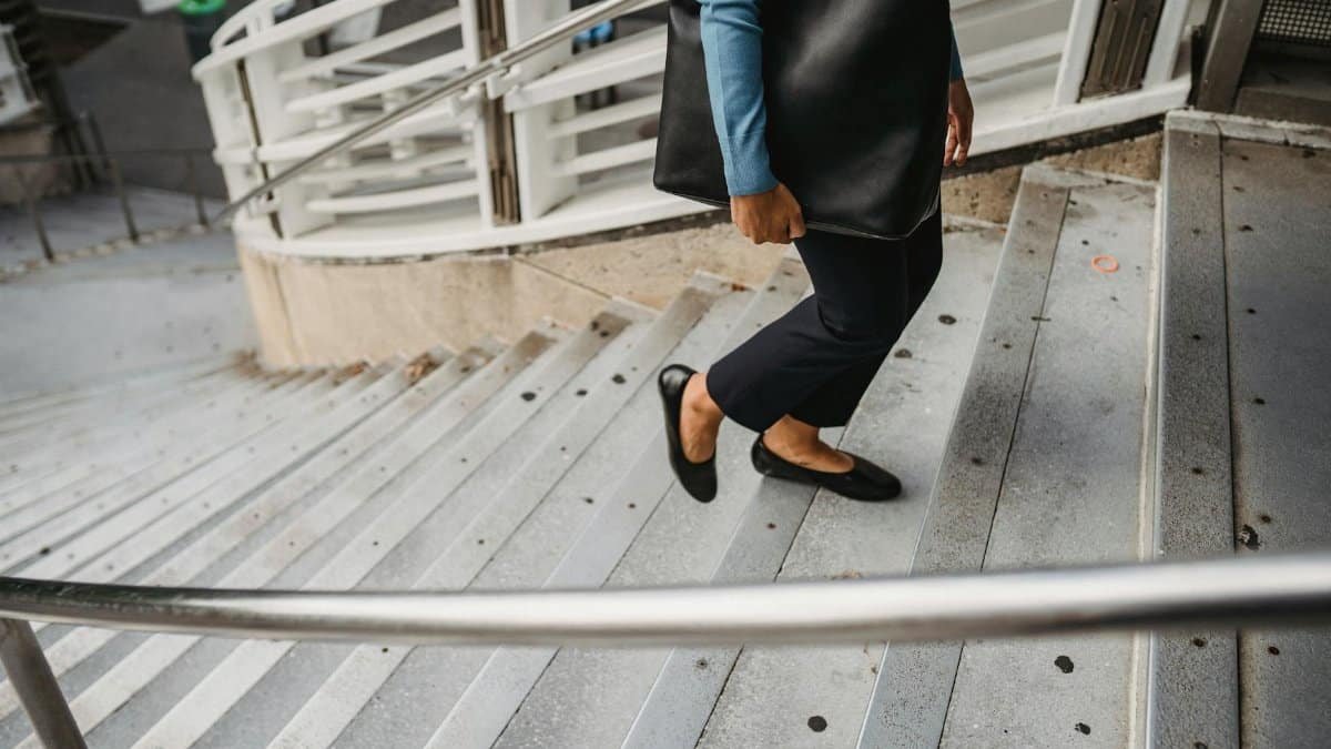 Unrecognizable female in casual clothes and black leather bag walking up concrete staircase with metal railing on street in city