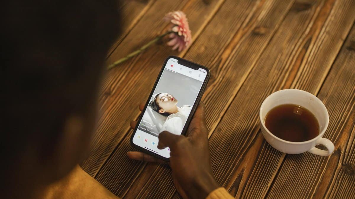 An individual using a dating app on a smartphone with a cup of tea nearby on a wooden table.