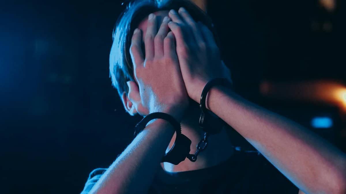 A person in handcuffs conceals their face under a blue light, suggesting a crime scene.