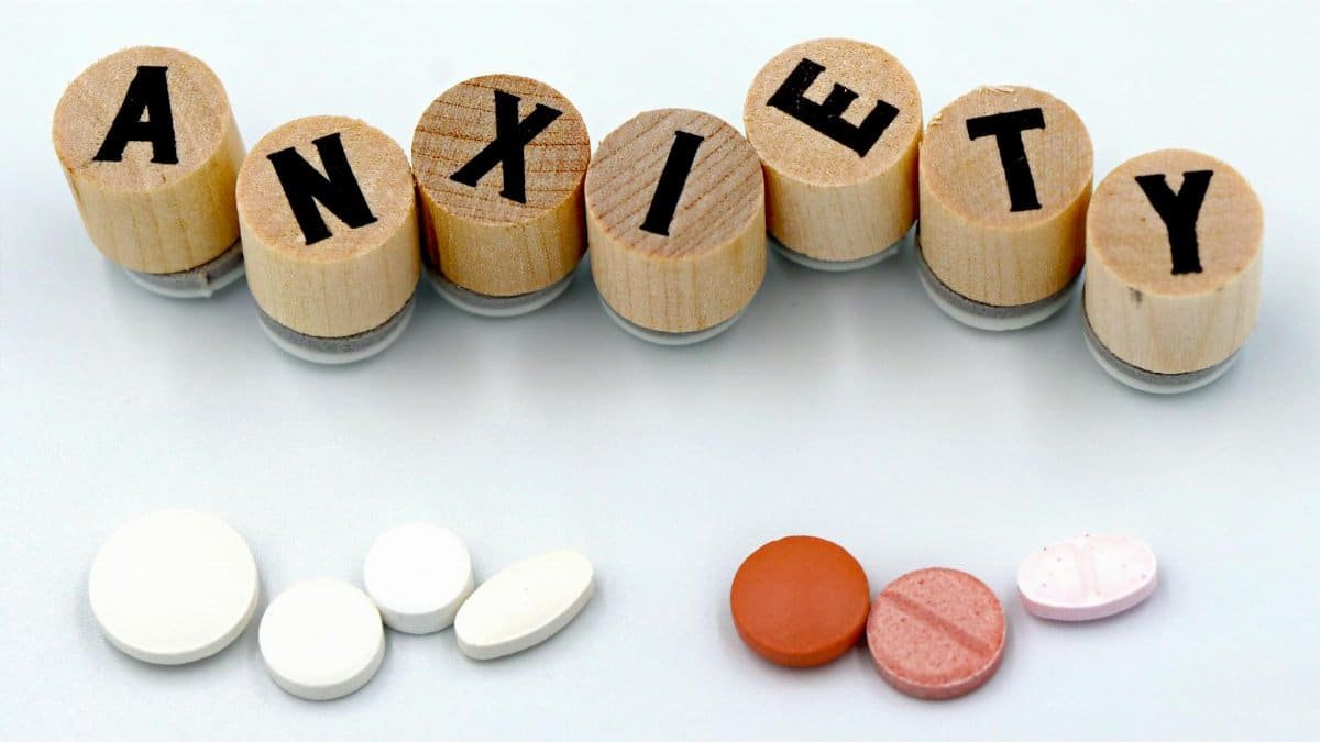 Wooden blocks spelling 'Anxiety' with assorted pills below on a light background.
