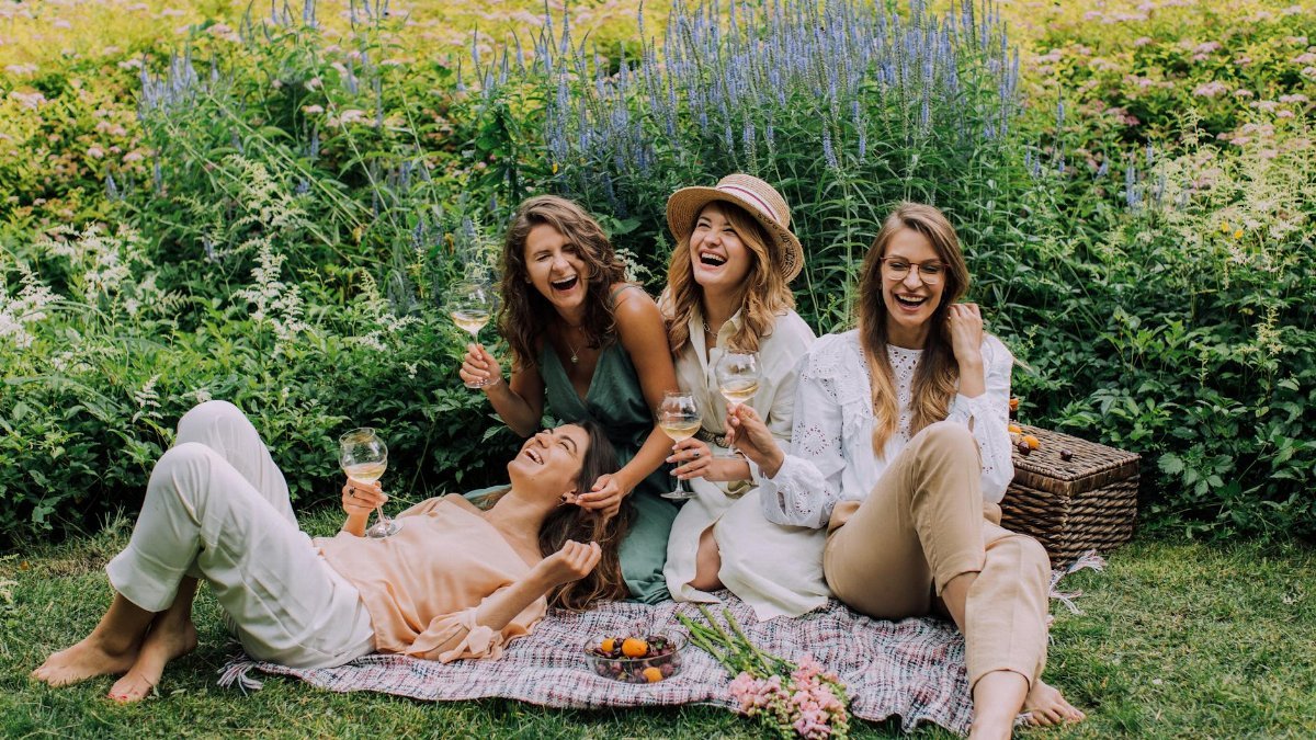 Four women enjoying a picnic, laughter, and wine in a vibrant summer garden.