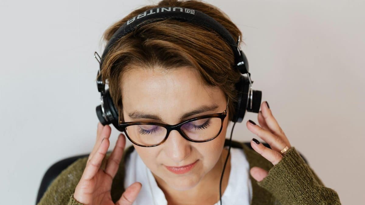 Positive relaxed female in casual warm jacket and eyeglasses resting against white wall with eyes closed and enjoying favorite music in wired headphones