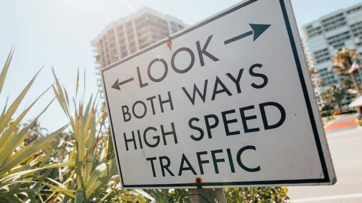 Direction sign with arrow and look both ways high speed traffic text placed on sidewalk near tropical plants against modern city buildings