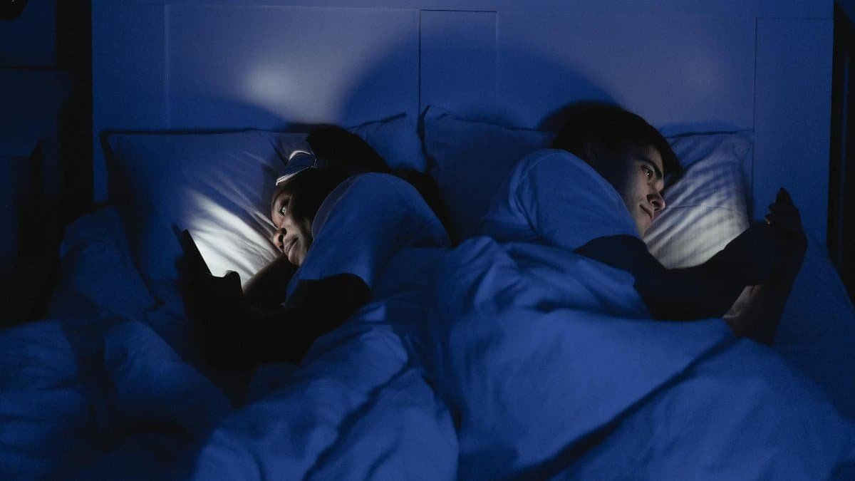 A couple lying in bed using smartphones at night, illuminated by screen light.