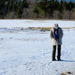 A lone hiker stands on a snow-covered field, surrounded by forest, capturing the essence of winter solitude.