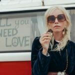 Fashionable woman in front of a retro van with 'All you need is love' sign, capturing the hippie vibe.