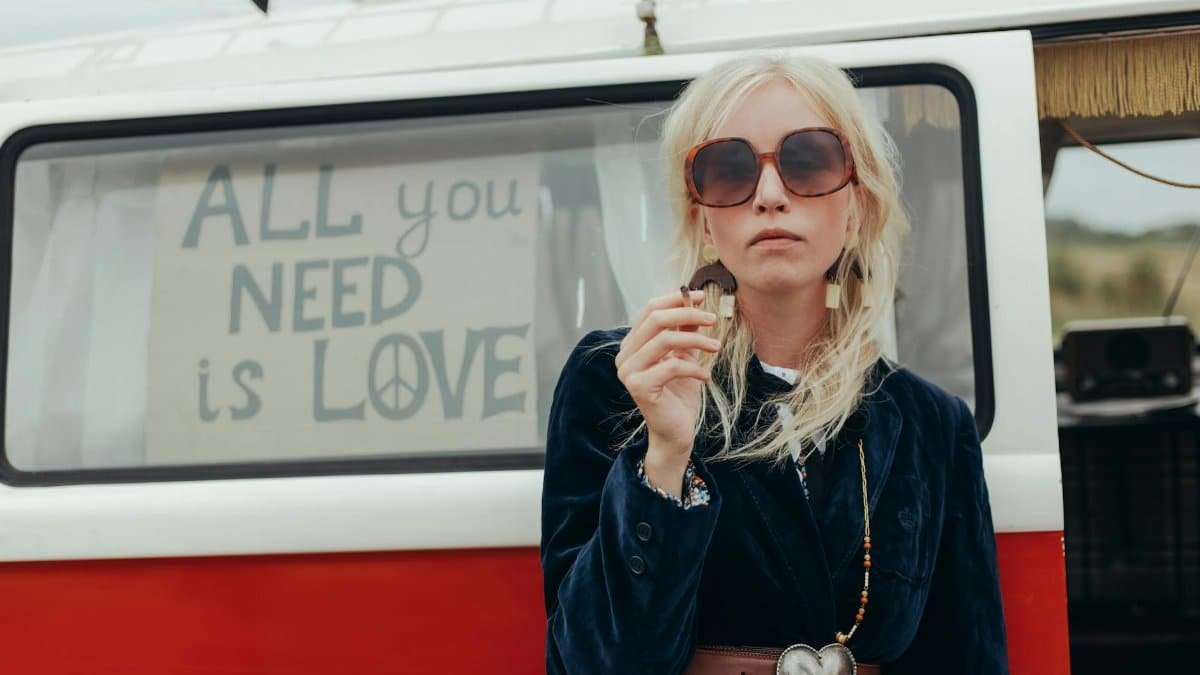 Fashionable woman in front of a retro van with 'All you need is love' sign, capturing the hippie vibe.