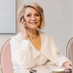 A smiling senior woman in a white blouse talking on the phone while working in a modern office environment.