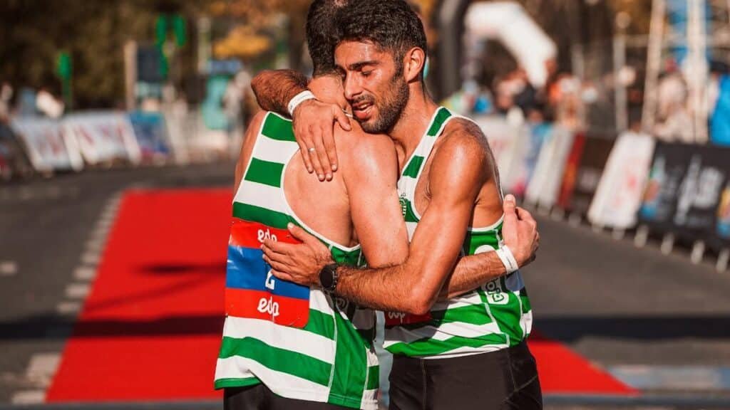 Two marathon runners in striped vests embrace at the finish line, celebrating sportsmanship.