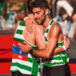 Two marathon runners in striped vests embrace at the finish line, celebrating sportsmanship.