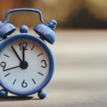 A classic blue alarm clock placed outdoors, displaying five minutes to twelve.