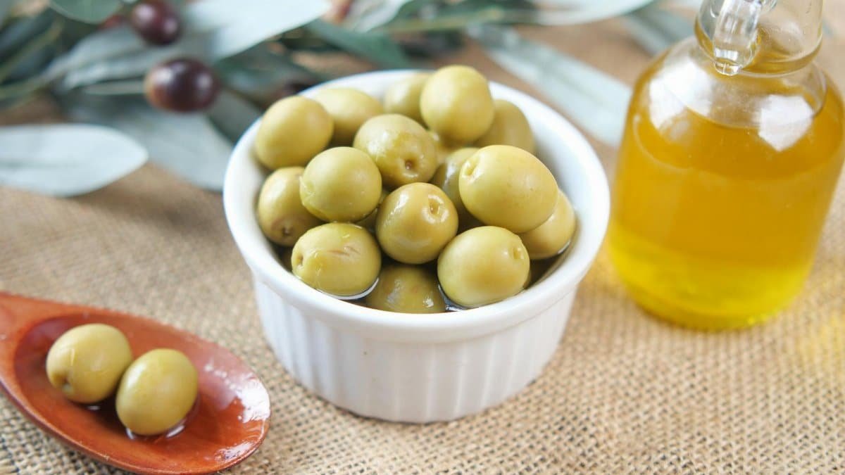 Close-up of fresh green olives in a bowl with a bottle of olive oil on a textured surface.