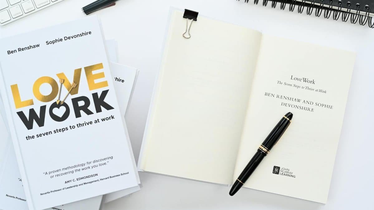 Flat lay of 'Love Work' books, open notebook, and pen on white desk, ideal for work themes.