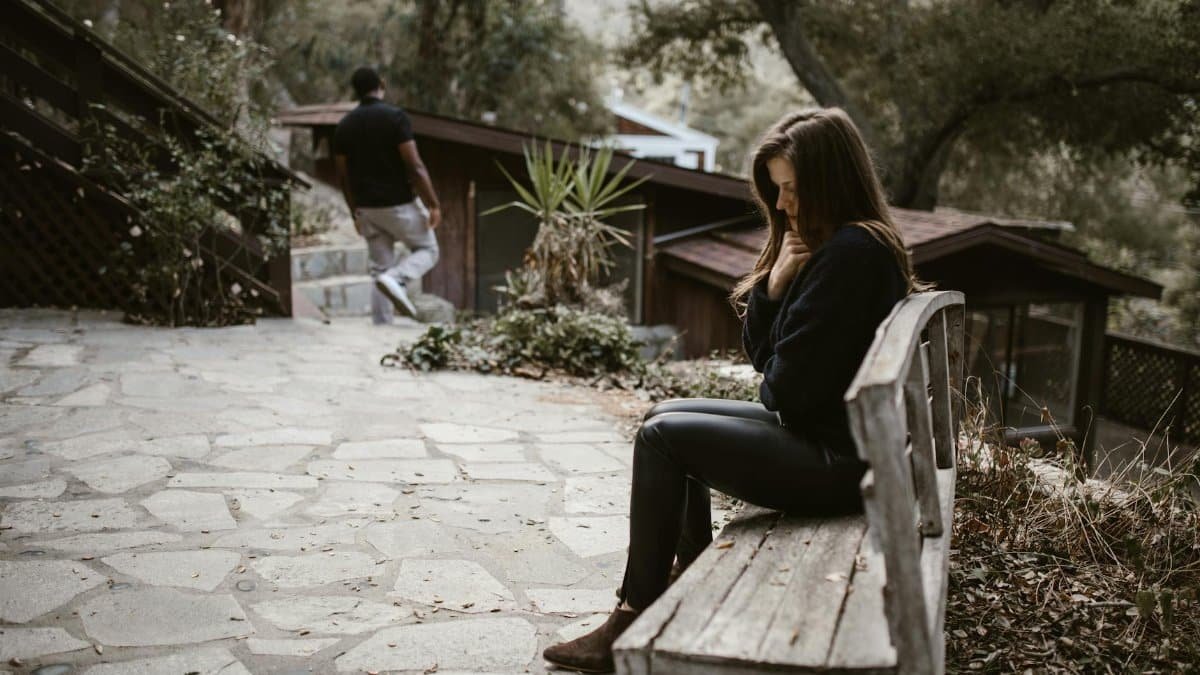 A woman sits pensively on a bench outside after a breakup, while a man walks away.