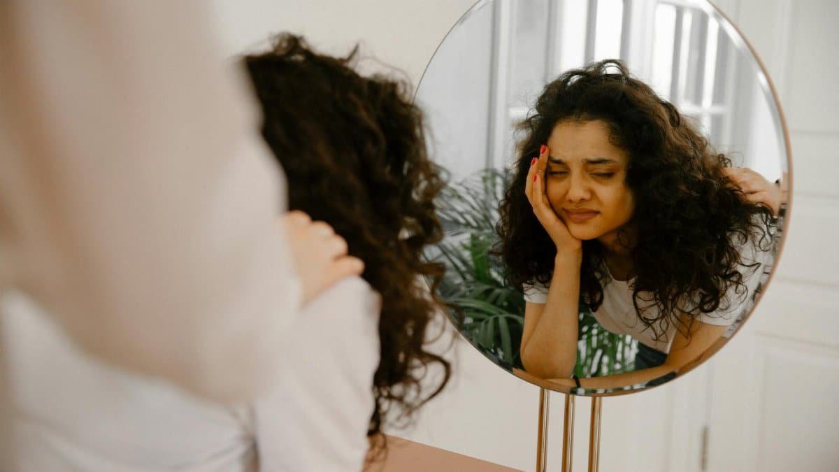 A young woman in front of a mirror expressing self-doubt and contemplation indoors.