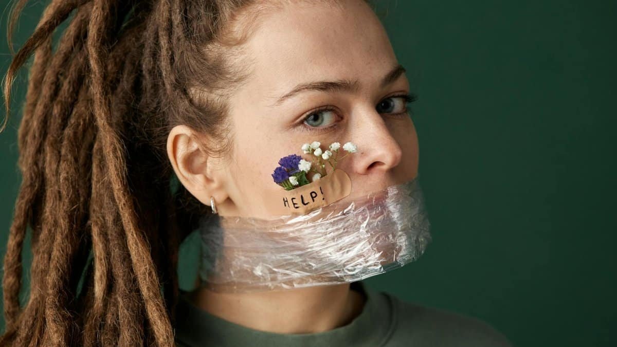 Creative portrait with woman’s mouth covered in plastic, flowers in band aid, and help message.