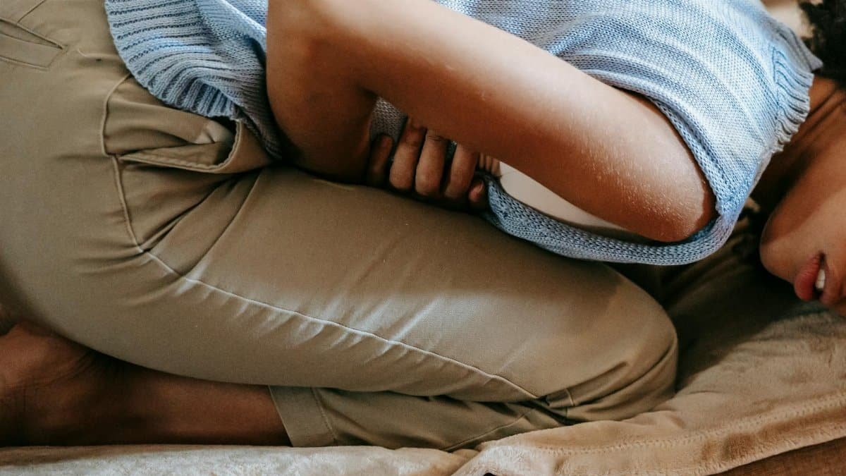 A young woman in discomfort, holding her stomach while curled up on a couch indoors.