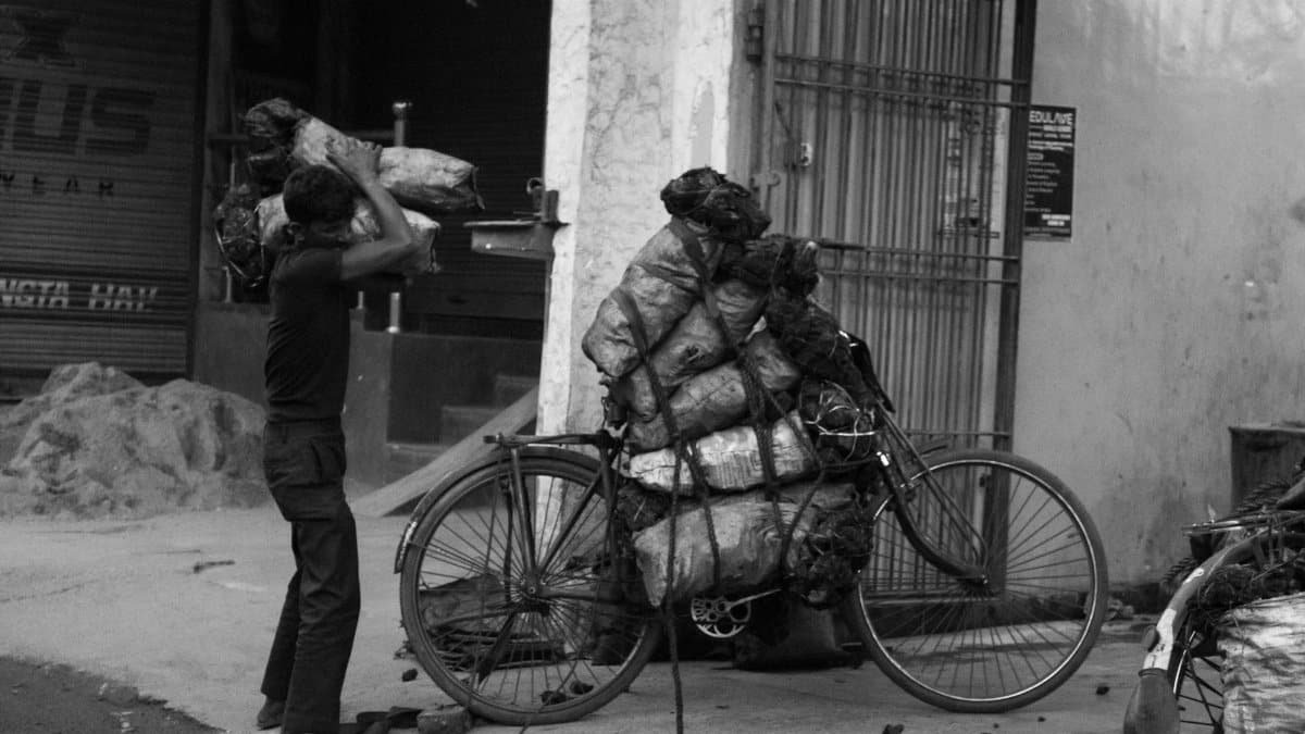 A person loads heavy bags onto a bicycle in an urban street, depicting hard work and manual labor.