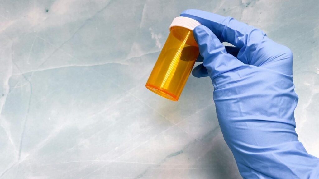 A gloved hand holding an empty orange pill bottle against a marble background.