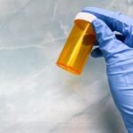 A gloved hand holding an empty orange pill bottle against a marble background.