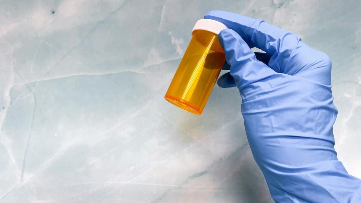 A gloved hand holding an empty orange pill bottle against a marble background.