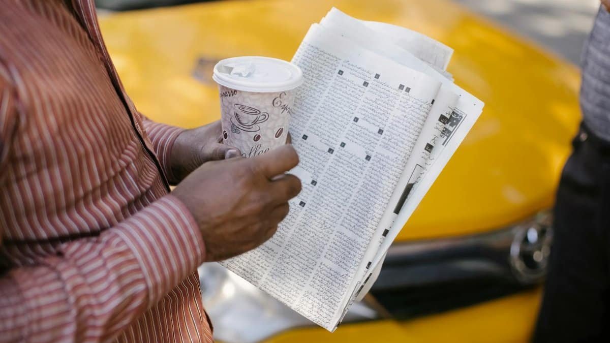 From above side view of crop anonymous ethnic male cab driver with coffee to go and newspaper talking to coworker on street