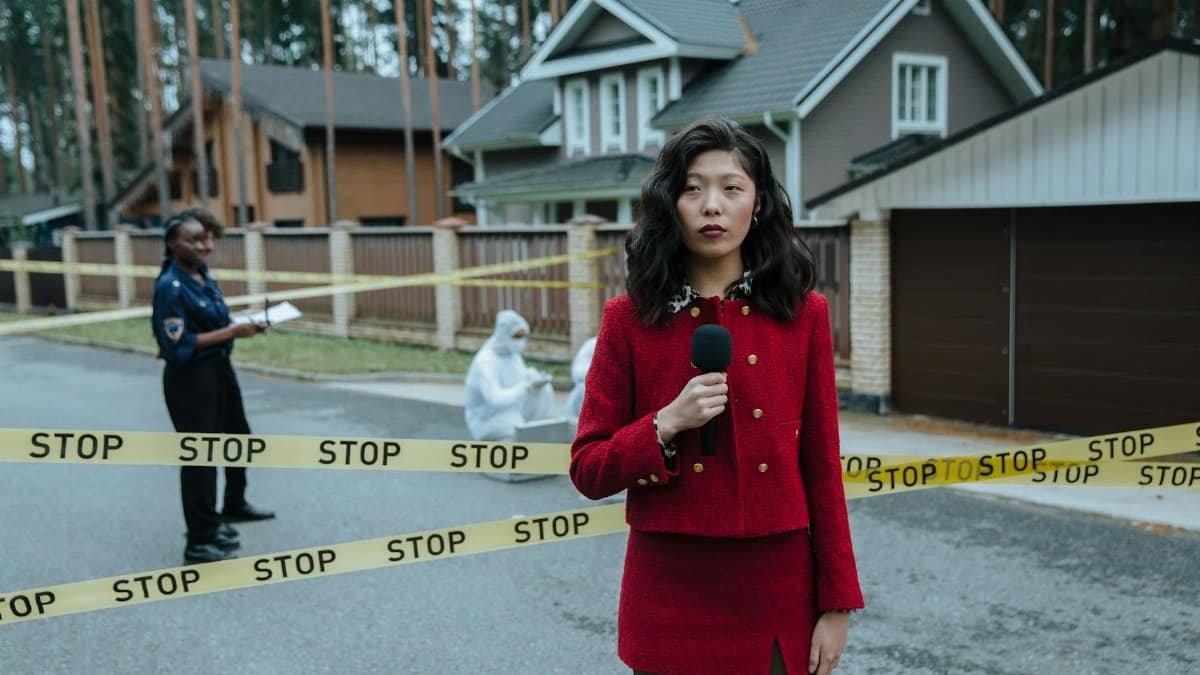 A journalist in a red outfit reporting live from a crime scene outdoors with stop tape and police presence.