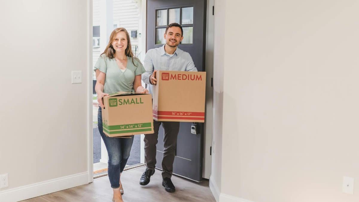 A smiling couple carrying cardboard boxes as they move into their new home.