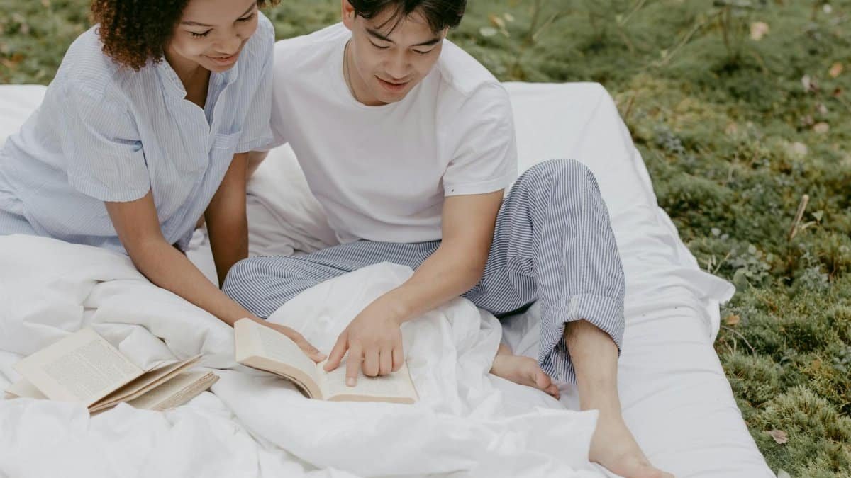 Couple enjoying a relaxing morning reading together on a white bed linen outdoors.