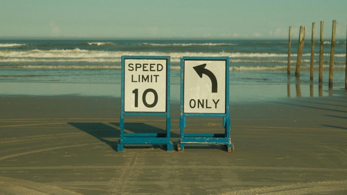 Beach scene with speed limit and left turn signs on the sand, ocean backdrop.