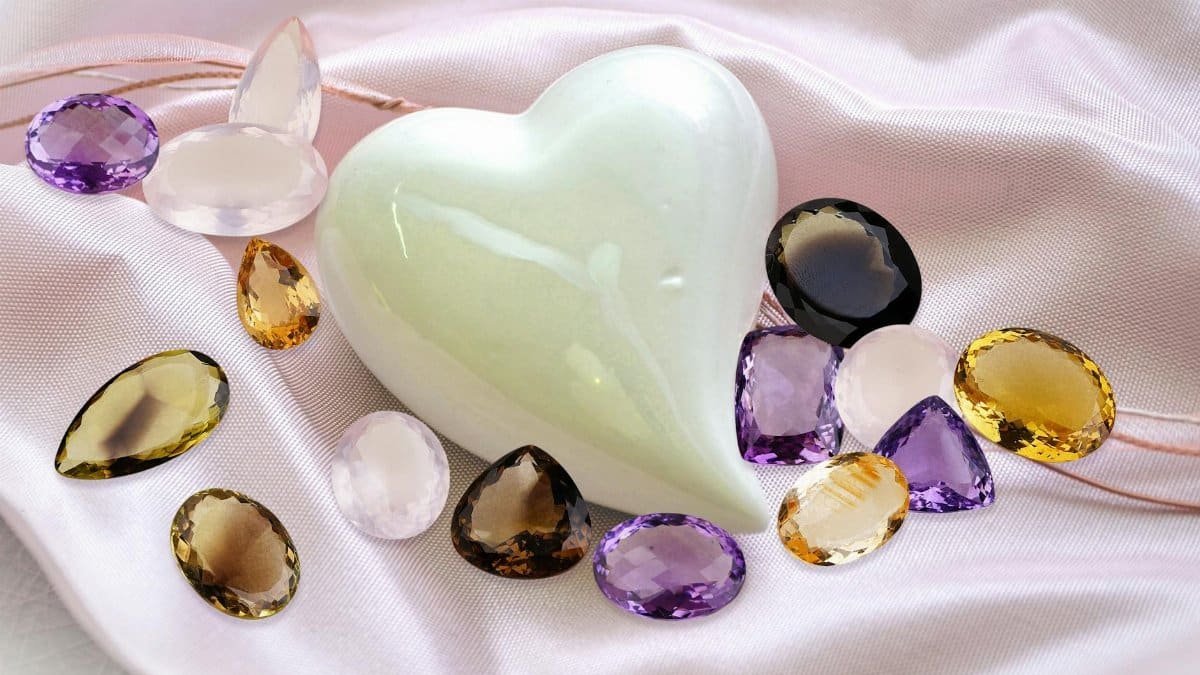 Heart-shaped jade surrounded by colorful gems on pink silk fabric. A beautiful and elegant close-up composition.