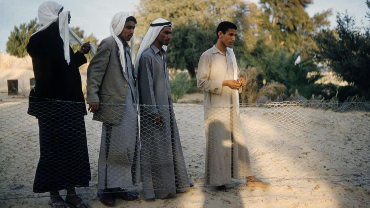 Group of Middle Eastern men in traditional attire beside a chain-link fence in a rural setting.