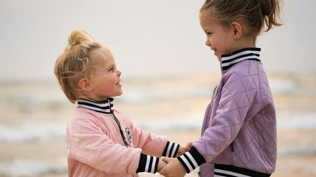 Two young girls smiling and holding hands on a beach, showcasing love and togetherness.