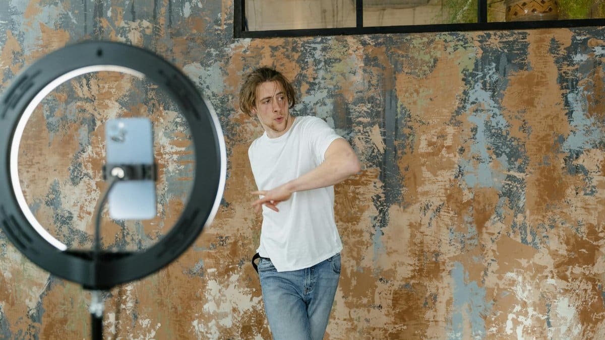 Young man in casual attire dances while recording a video indoors with a smartphone and ring light.