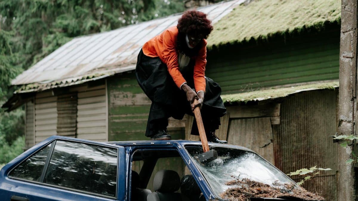 A creepy clown in costume with a sledgehammer aggressively smashing a car windshield outdoors.