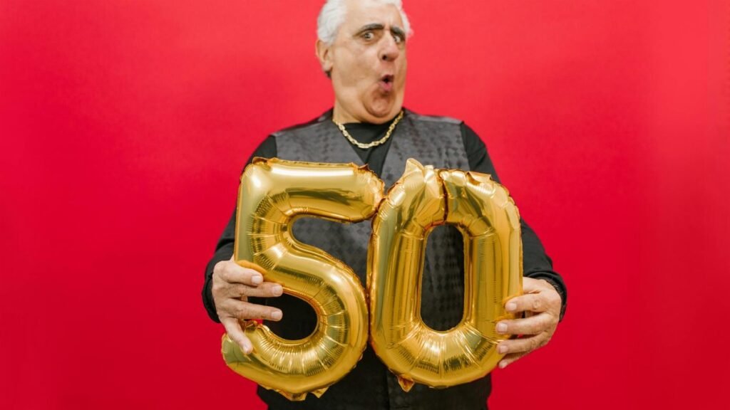 A joyful senior holding gold '50' balloons against a vibrant red backdrop.