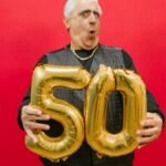 A joyful senior holding gold '50' balloons against a vibrant red backdrop.