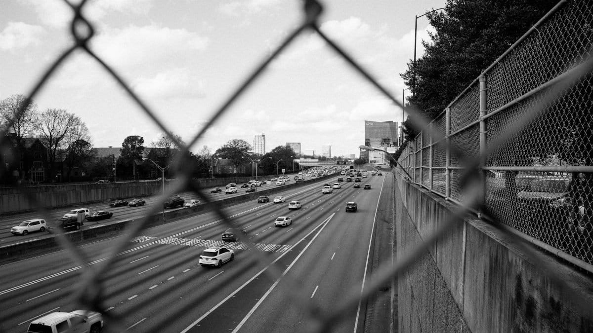 A black and white photo of a busy urban highway seen through a chain-link fence.