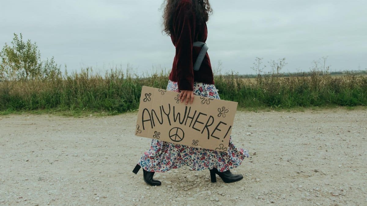 Side view of a hippie woman holding an 'Anywhere' sign on an unpaved road.