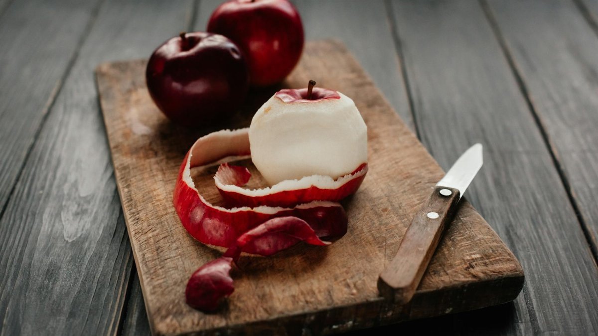 A peeled red apple with a knife on a rustic wooden chopping board.