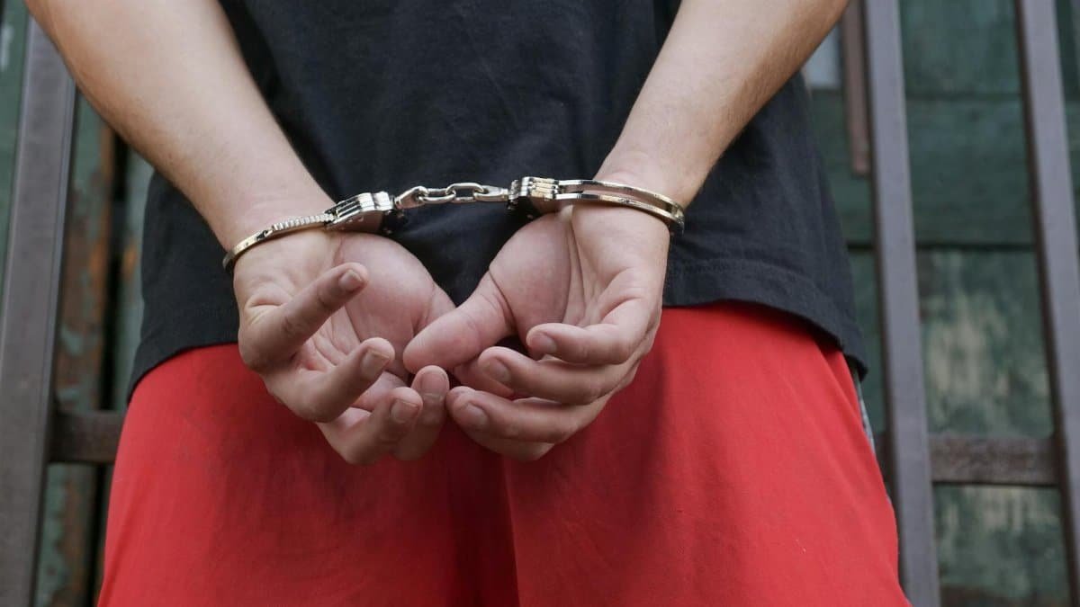 Close-up of a person in handcuffs, viewed from behind, wearing red pants and a black shirt.