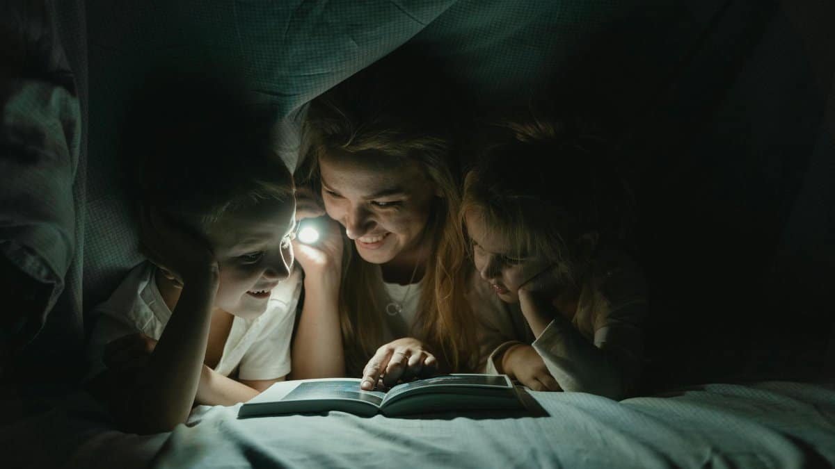 A mother reads a bedtime story to her children under the blanket with a flashlight, creating a warm, bonding moment.