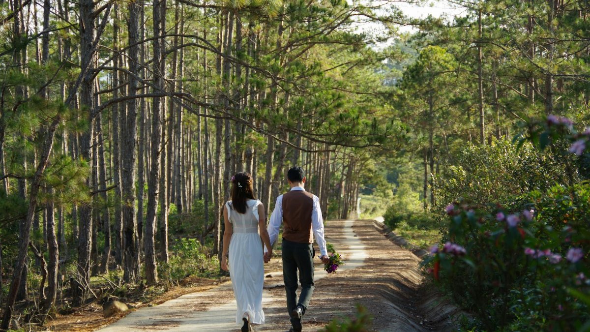 A couple walks hand-in-hand down a serene forest path, surrounded by lush greenery.