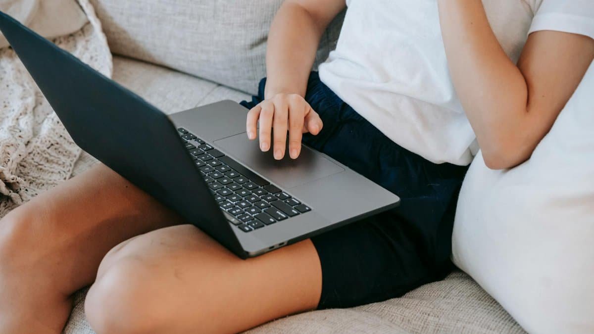 From above of crop unrecognizable self employed young lady in casual clothes sitting on comfortable couch and working online on laptop at home