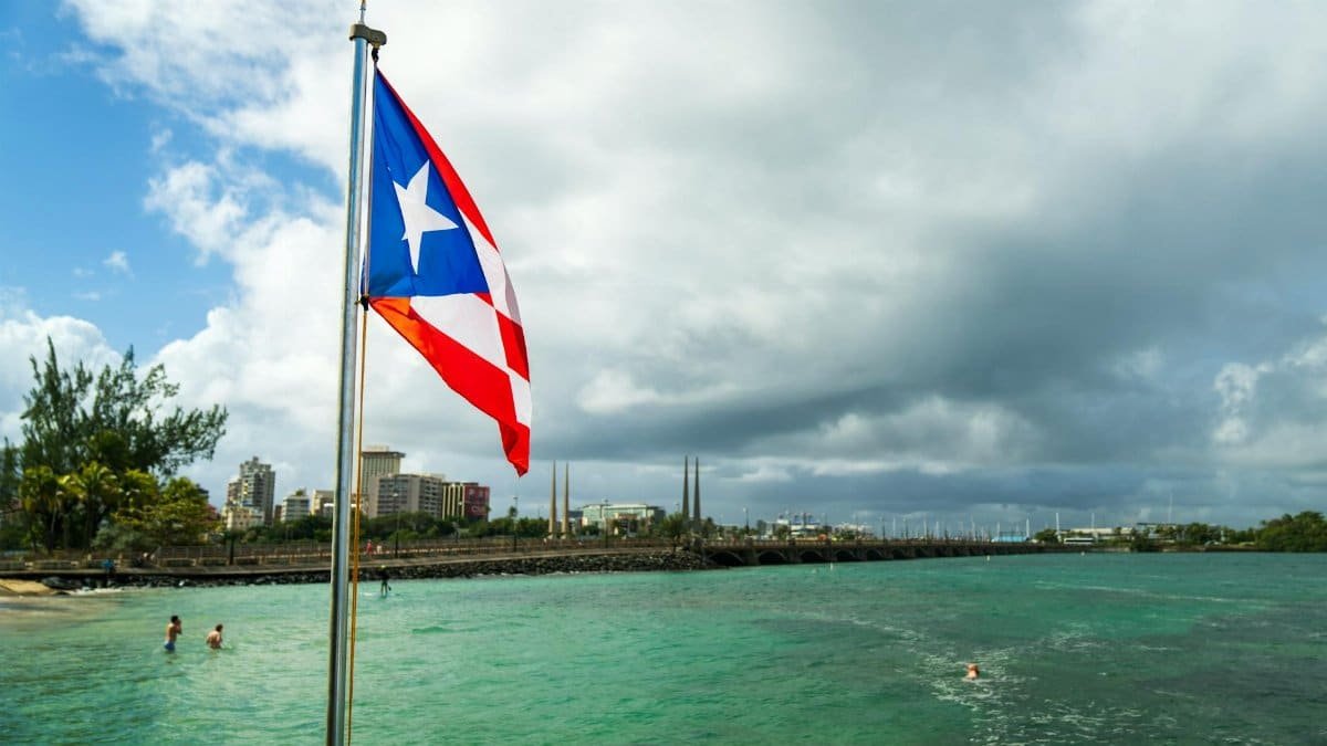 Puerto Rican flag on a flagpole by the sea with city skyline in the background.
