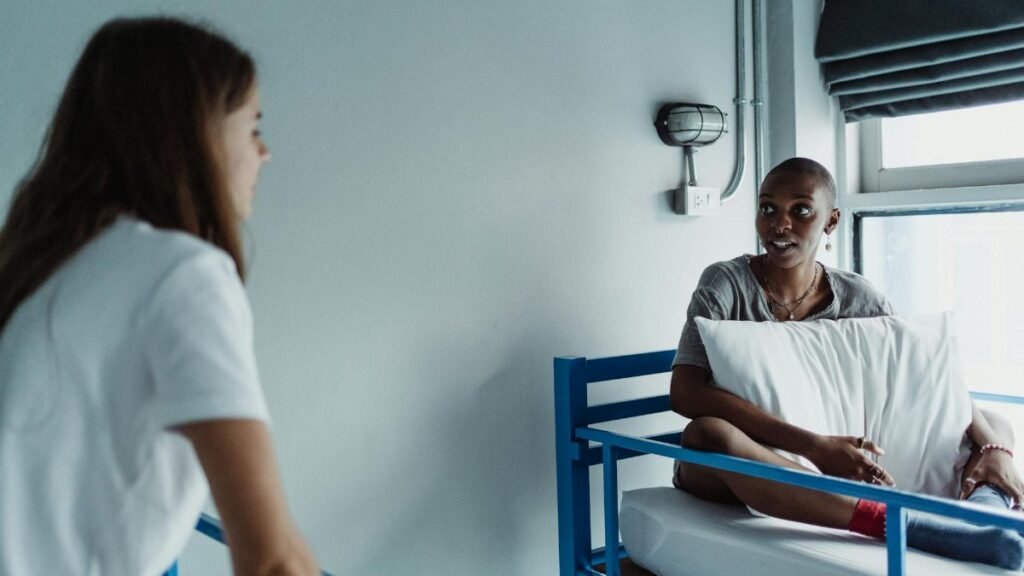 Two women talking in a hospital room, one sitting on a bed holding a pillow.