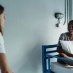 Two women talking in a hospital room, one sitting on a bed holding a pillow.