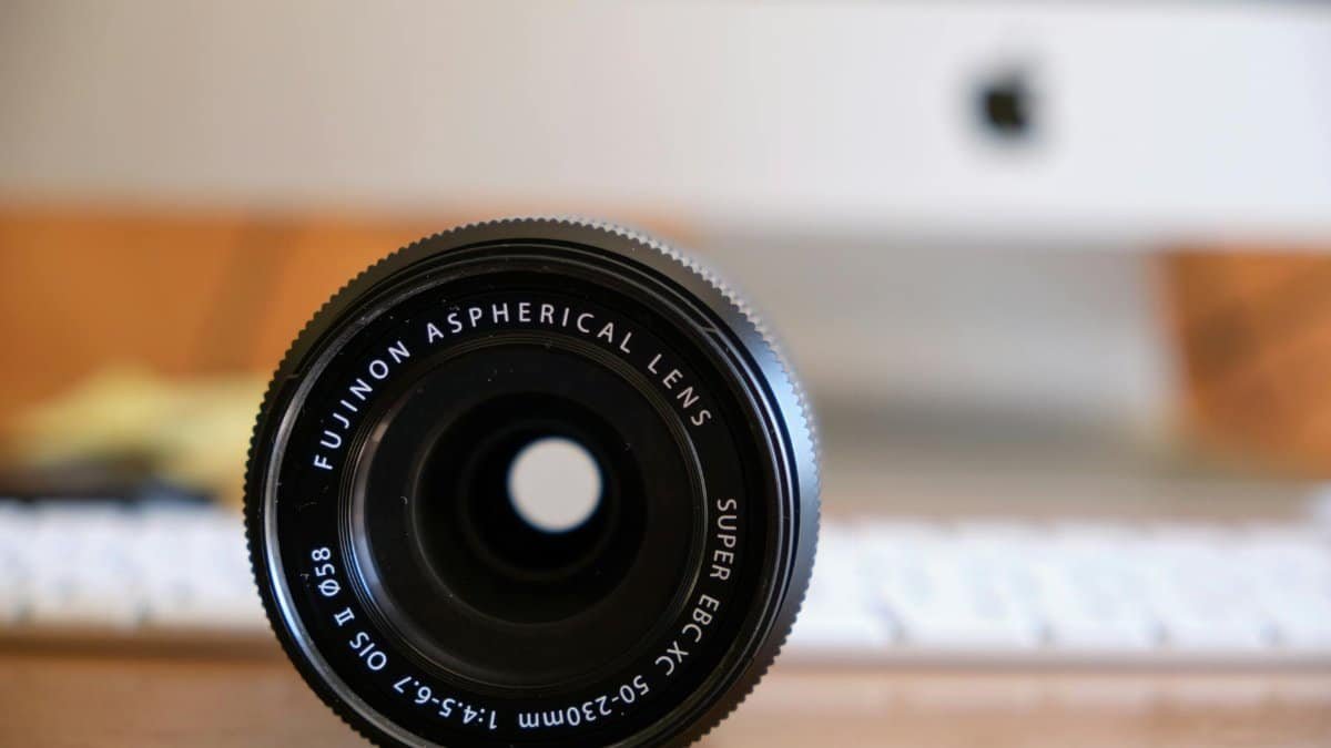 Close-up of a Fujinon aspherical lens with a blurred computer in the background, on a wooden desk.