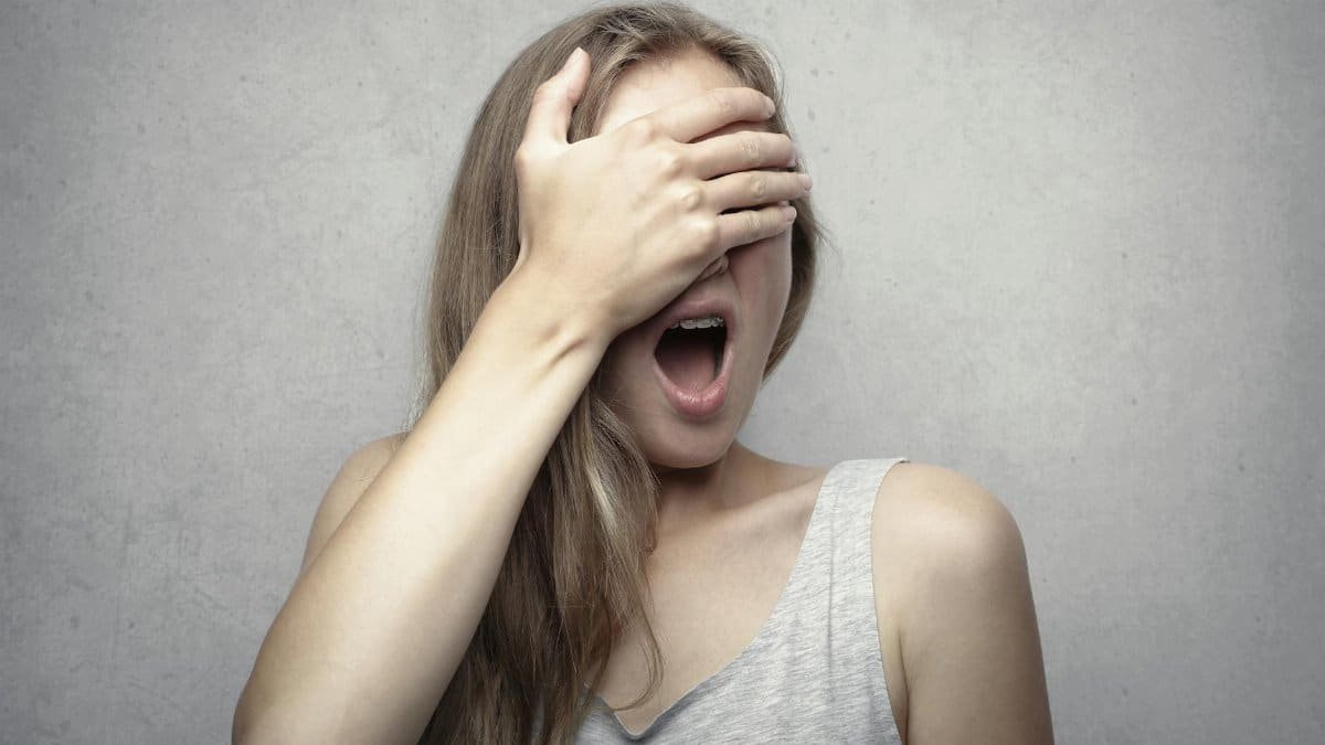 A surprised woman covers her face with one hand, expressing shock and amazement against a gray background.