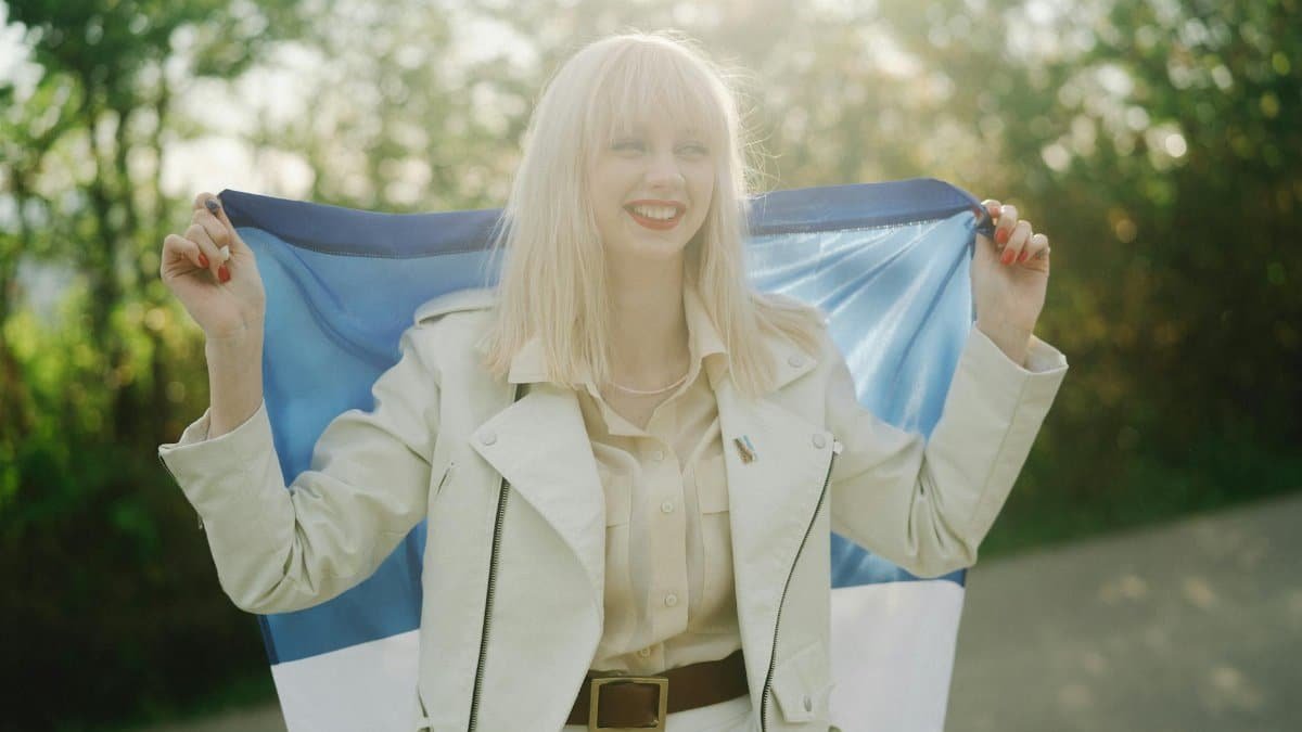 Blonde woman in beige coat joyfully holding a flag in a sunny outdoor setting.